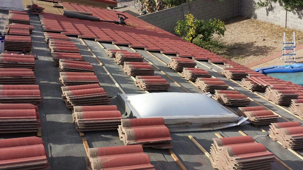 Roof in mid-installation showing red clay tiles stacked and partially laid on underlayment, with a skylight visible in the center of the work area.