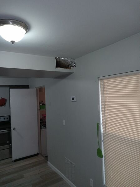 Interior room view showing an exposed flexible duct opening in the wall near the ceiling, along with a thermostat, floor vent, and kitchen area in the background.