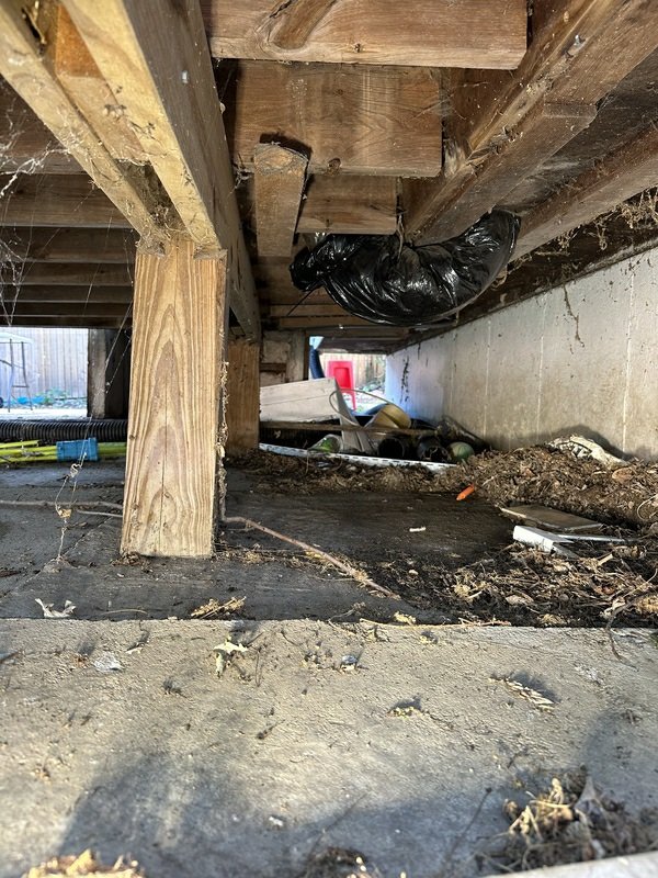 Under-deck/crawl space area showing debris, trash bags, dirt accumulation, and corrugated ducting — relevant to HVAC condition documentation.