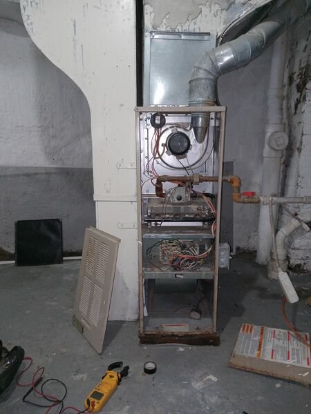 An open furnace unit with panels removed, exposing internal components including the blower, heat exchanger, and wiring in a basement utility area.