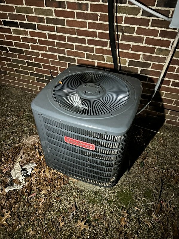Exterior Goodman HVAC condenser unit photographed at night against a brick wall, clearly showing the full unit in context.