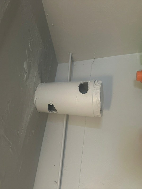 A white HVAC duct/pipe section mounted at the wall-ceiling junction showing two significant holes punched through it, clearly documenting damage or a defective condition.