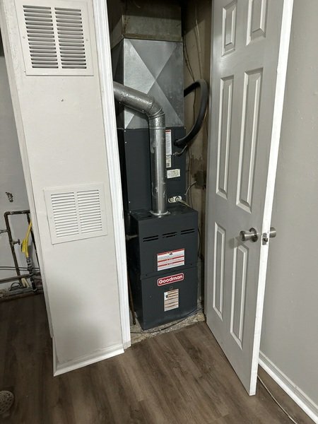 A Goodman furnace unit visible inside an open utility closet, showing the indoor HVAC component with ductwork and exhaust piping.