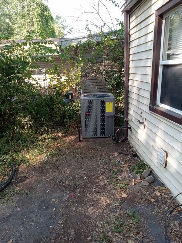 Exterior HVAC condenser unit installed beside the house, surrounded by overgrown vegetation and debris on bare dirt.
