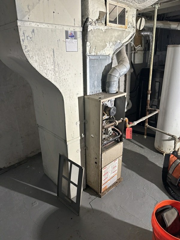 An older furnace unit with open panel, ductwork, and associated piping in a basement utility area, showing the HVAC system's condition.