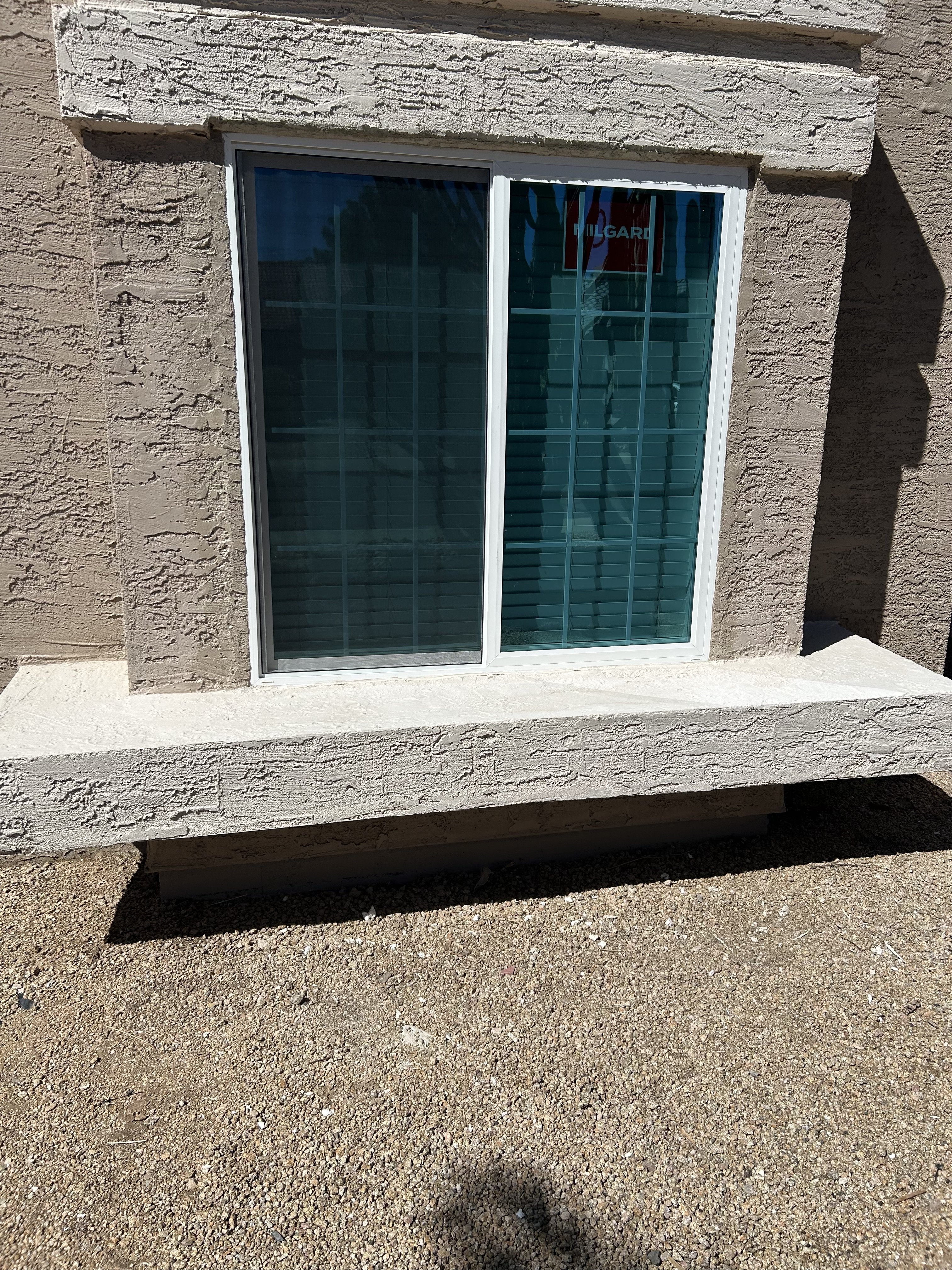 A newly installed Milgard sliding window set in a stucco exterior wall, shown from outside with a concrete sill and gravel ground below.