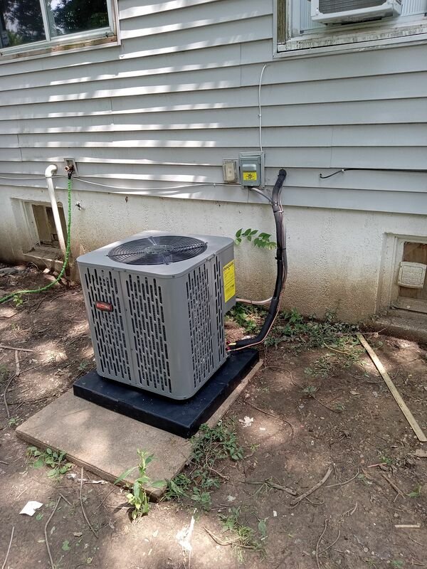 Exterior central air conditioning condenser unit mounted on a pad beside the house, with refrigerant lines and electrical disconnect visible.