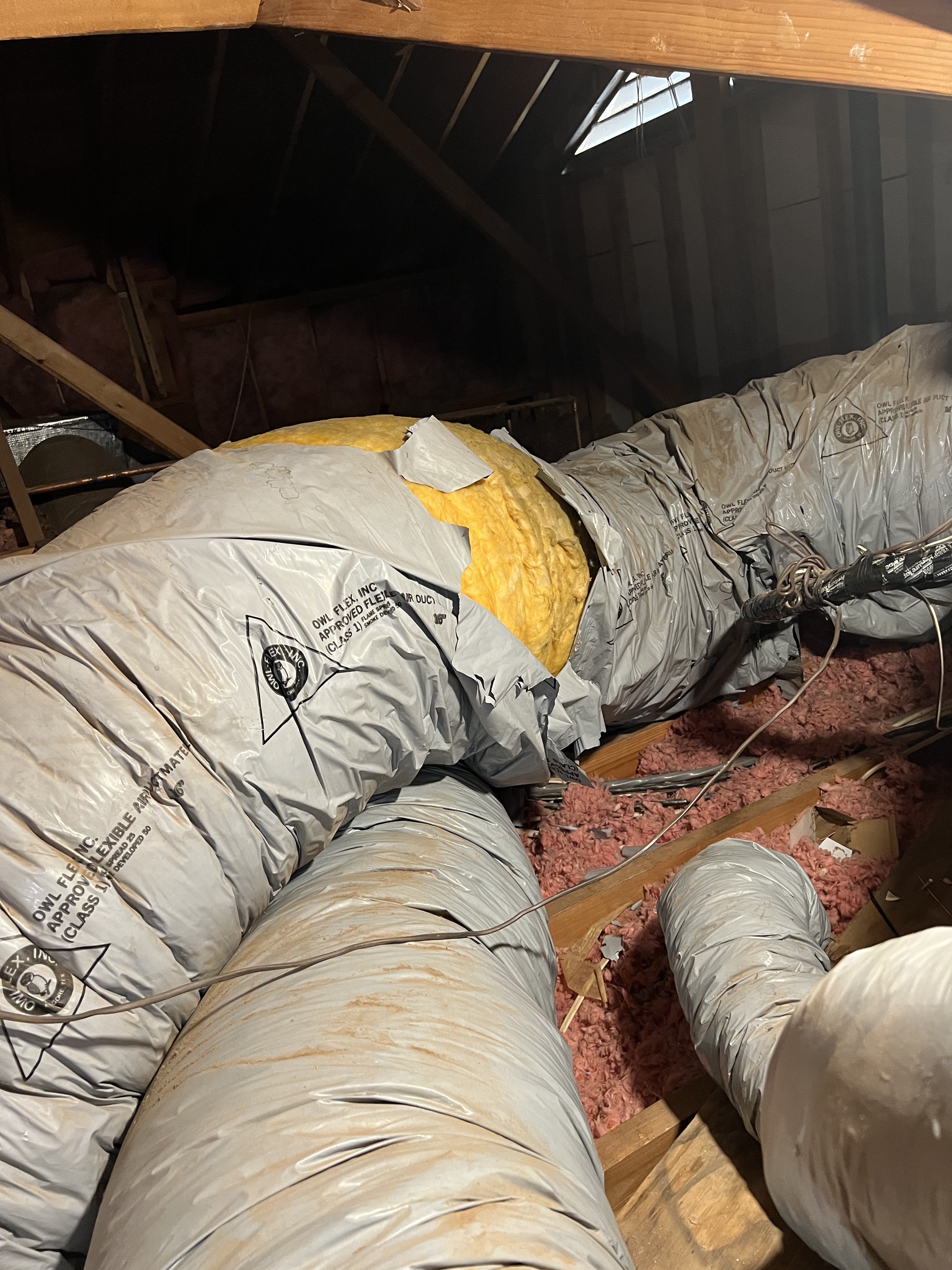 Attic interior showing flexible HVAC ductwork (Owl Flex brand) with visible damage — outer insulation jacket torn open exposing yellow fiberglass insulation — alongside pink blown-in insulation on the attic floor.