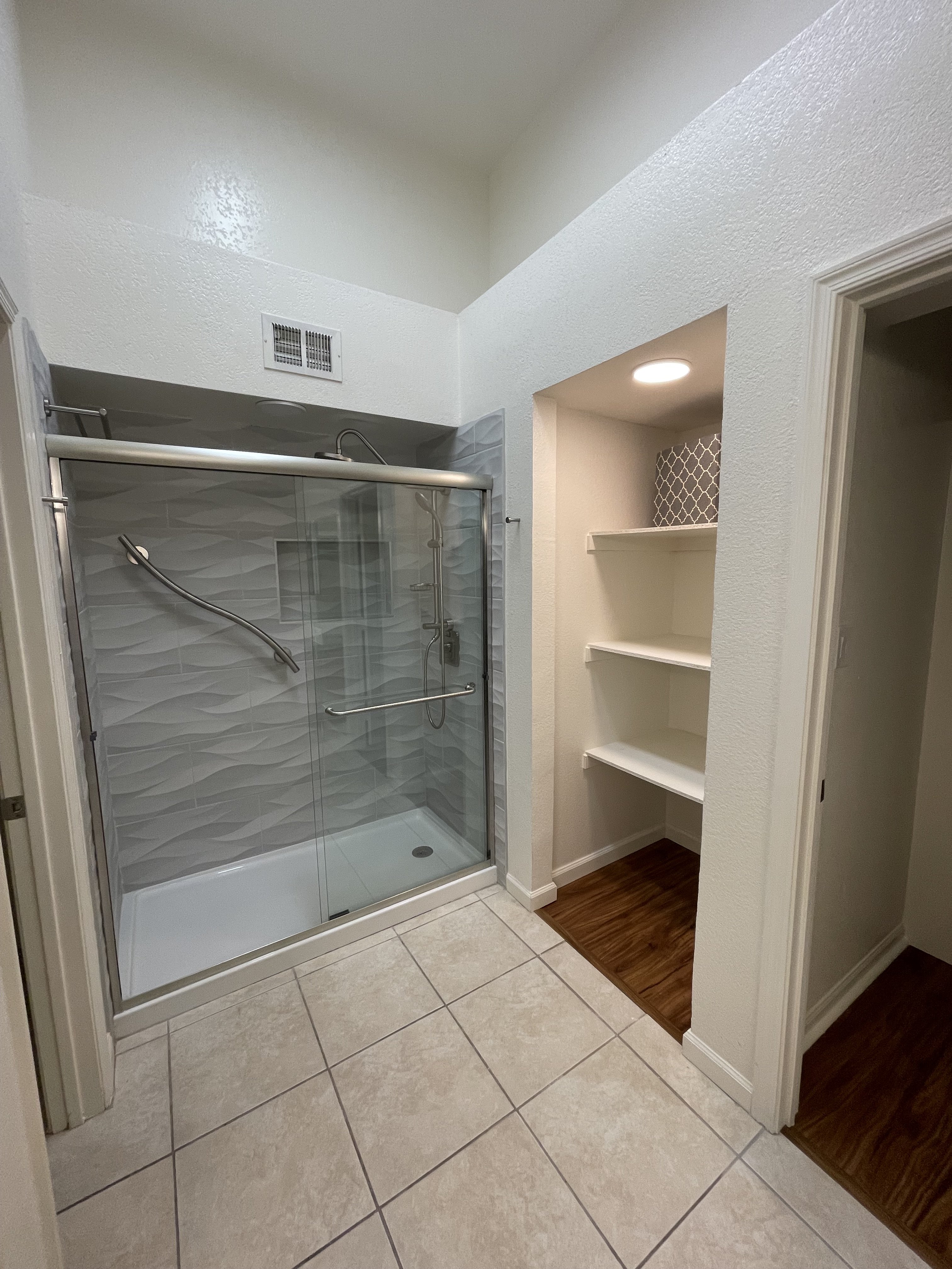 A clean, well-lit bathroom showing a walk-in shower with glass sliding doors, wave-tile surround, grab bar, and handheld shower fixtures, alongside a built-in closet with shelving — relevant to plumbing scope.