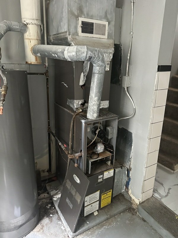 A furnace/HVAC unit with ductwork, open access panel, and visible deterioration in a utility room, clearly showing the system's condition.