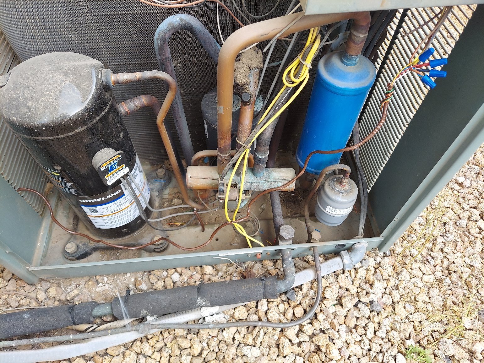 Interior view of an HVAC condenser unit showing a Copeland compressor, refrigerant lines, accumulator, and associated components with visible wear and corrosion.