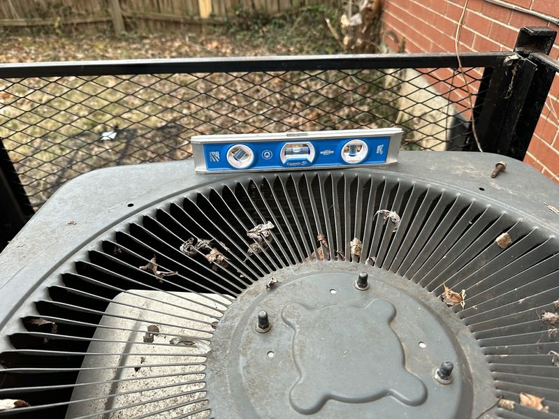 Top-down view of an outdoor AC condenser unit with a spirit level placed on it, showing debris accumulation and a level check being performed.